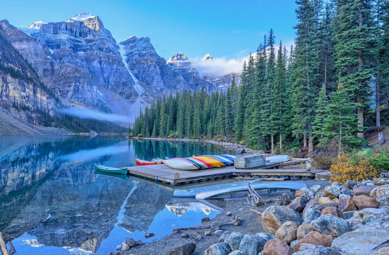 A still alpine lake with canoes on a dock below snow-capped peaks and pines